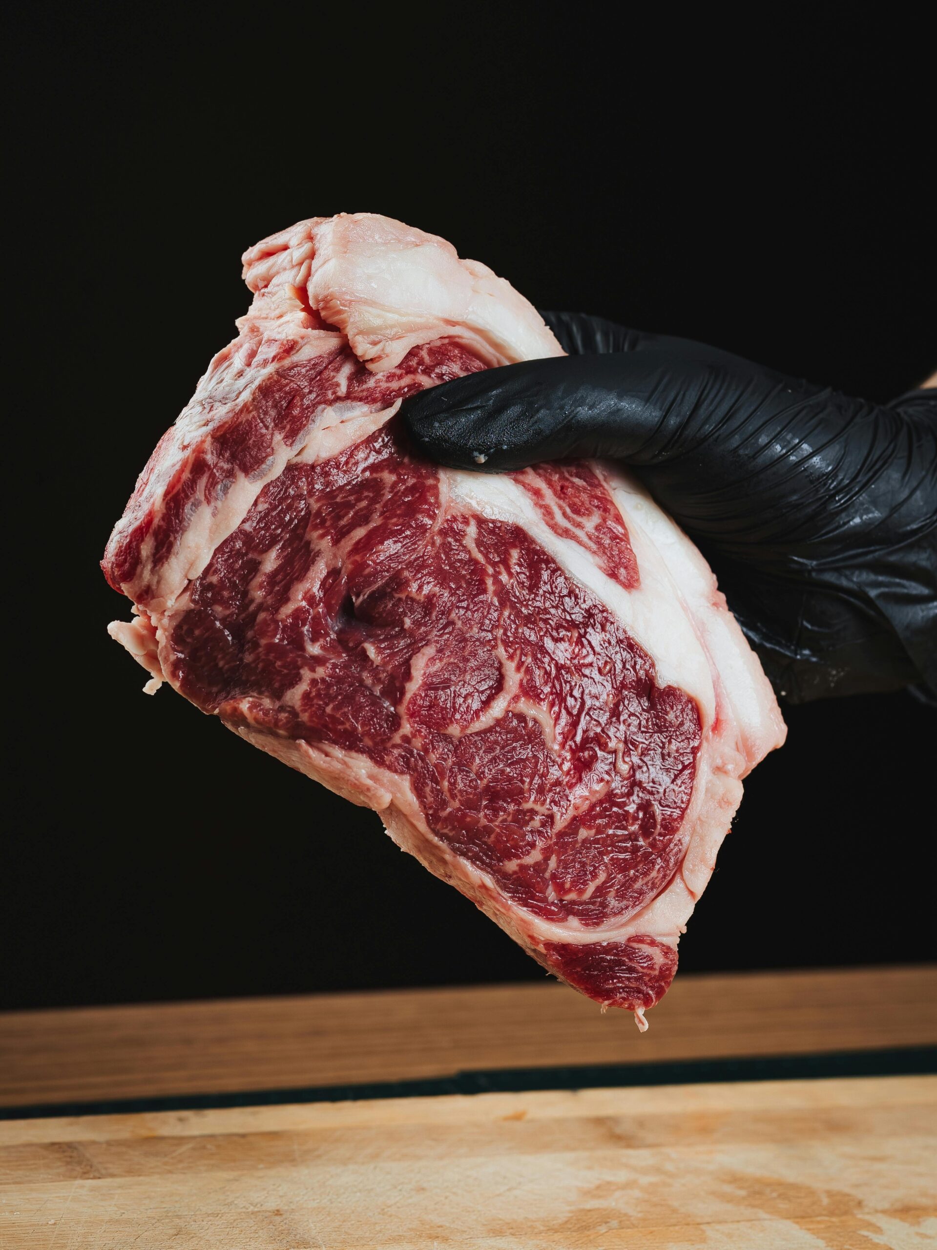 Close-up of a raw ribeye steak held by a gloved hand on a wooden surface.